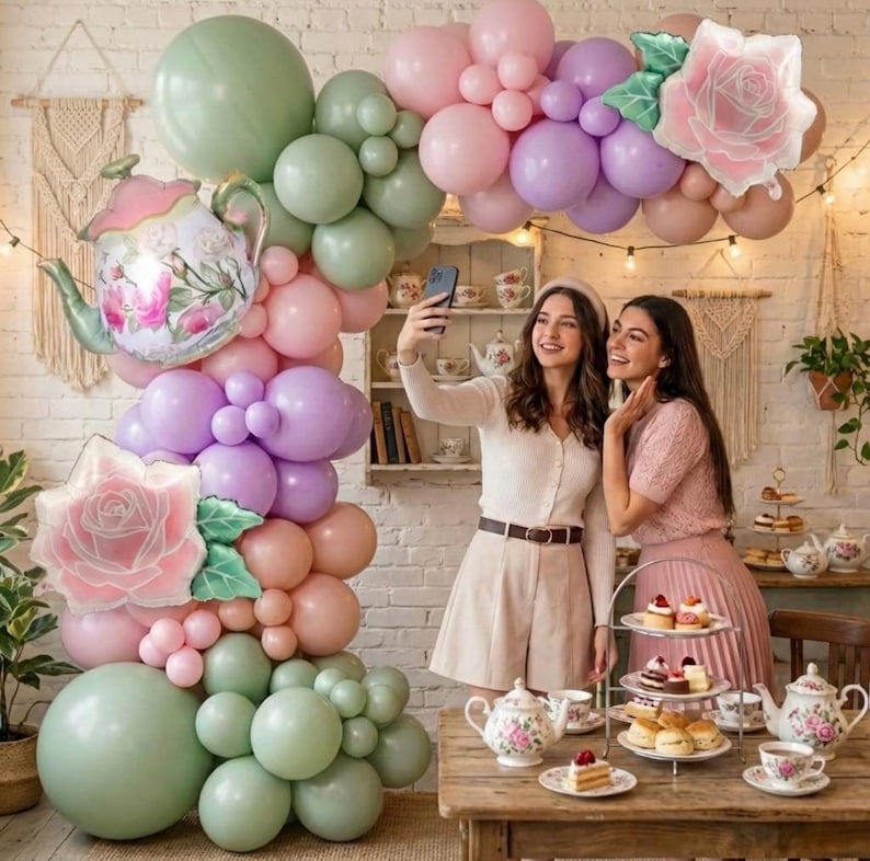 K&ouml;nnte beinhalten: Eine verspielte Party-Szene mit einem Ballonbogen in Pastellfarben, darunter Rosa, Lila und Gr&uuml;n. Ein Teekannen-Ballon und ein rosenf&ouml;rmiger Ballon erg&auml;nzen die Dekoration. Zwei Personen machen ein Selfie bei einer Teeparty.