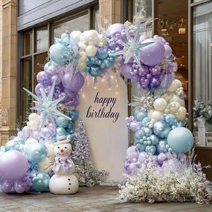 Puede incluir: Decoraci&oacute;n festiva de cumplea&ntilde;os con un arco de globos en tonos morados, azules y blancos. El arco enmarca un tel&oacute;n de fondo blanco con la frase "happy birthday". Una estatua de un mu&ntilde;eco de nieve est&aacute; al frente.