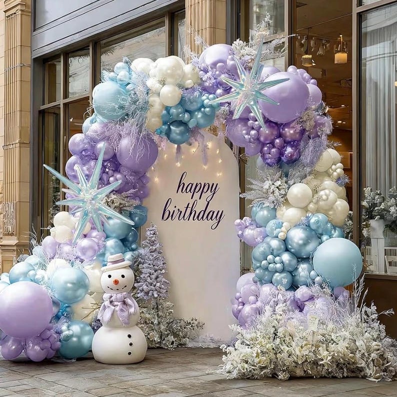 Puede incluir: Decoraci&oacute;n festiva de cumplea&ntilde;os con un arco de globos en tonos morados, azules y blancos. El arco enmarca un tel&oacute;n de fondo blanco con la frase "happy birthday". Una estatua de un mu&ntilde;eco de nieve est&aacute; al frente.