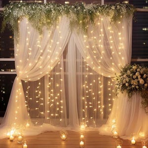 Peut inclure: Rideaux blancs transparents ornés de guirlandes lumineuses et de compositions florales. L'ensemble est éclairé par une lumière douce et chaleureuse, créant une ambiance romantique et élégante. Des bougies dans des photophores sont posées au sol.