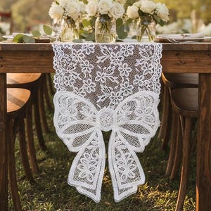 May include: A white lace table runner with a large bow design at the end. The runner is placed on a wooden table, with white flowers in glass jars in the background. The lace features floral patterns.
