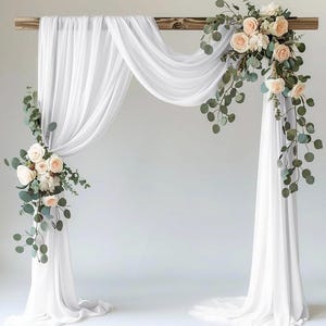 May include: A white draped wedding arch with a wooden top, adorned with cascading white fabric and floral arrangements of blush roses and eucalyptus. The arch is set against a plain white backdrop, creating an elegant and romantic setting.