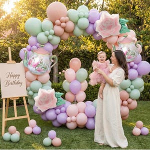 K&ouml;nnte beinhalten: Ein pastellfarbener Ballonbogen mit floralen Teekannen- und Rosendekorationen. Eine h&ouml;lzerne Staffelei zeigt ein Schild mit der Aufschrift "Happy Birthday". Eine Frau h&auml;lt ein Baby in einem rosa Kleid.