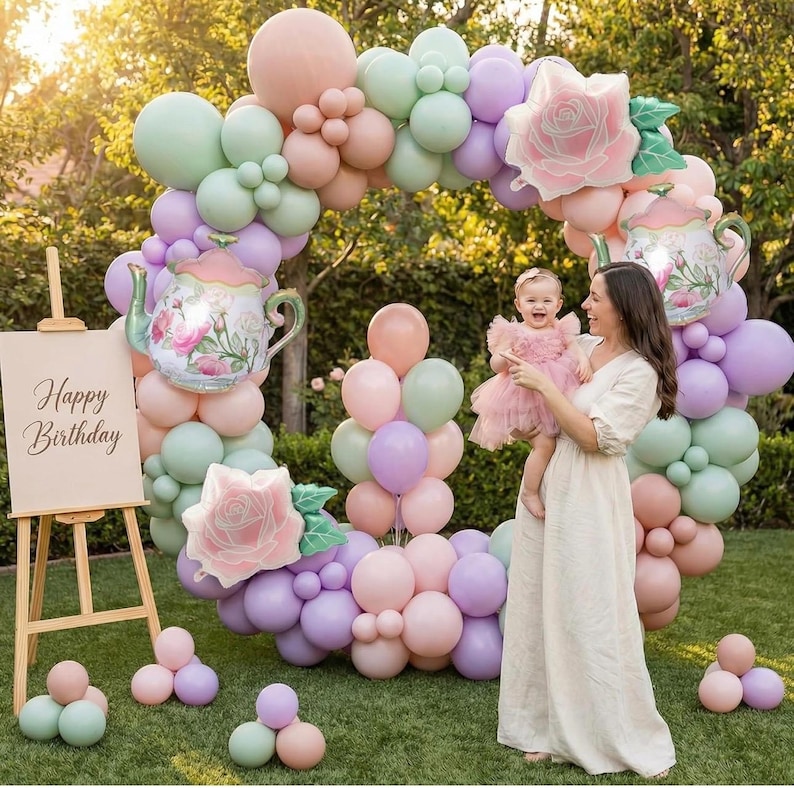 K&ouml;nnte beinhalten: Ein pastellfarbener Ballonbogen mit floralen Teekannen- und Rosendekorationen. Eine h&ouml;lzerne Staffelei zeigt ein Schild mit der Aufschrift "Happy Birthday". Eine Frau h&auml;lt ein Baby in einem rosa Kleid.