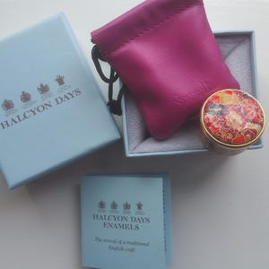 以下が含まれることがあります： 「HALCYON DAYS」と「ENAMELS」の文字が入った水色の箱と小冊子。 金色の縁取りと花柄模様の小さな丸い小物入れが、水色の箱の中のマゼンタ色のポーチの隣に置かれています。