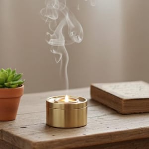 Op de afbeelding: Een brandende gouden kaars in een ronde houder met opstijgende rook, een kleine potplant en een gesloten boek op een houten tafel. De kaars staat in het midden, met de rook die omhoog dwarrelt.