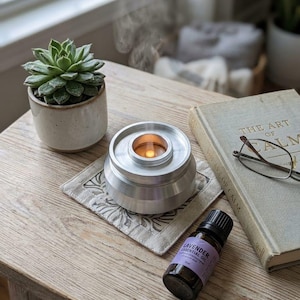 Op de afbeelding: Een zilveren aromatherapie diffuser met een brandende kaars, die stoom afgeeft, staat op een onderzetter met patroon. Een vetplant in een keramische pot, een boek getiteld "The Art of Calm" en een flesje lavendel etherische olie liggen op een houten tafel.