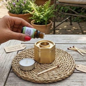 Puede incluir: Un difusor de aceites esenciales hexagonal dorado con una vela y cerillas sobre una esterilla tejida. Una mano vierte aceite de una botella etiquetada "Lavender & Fern Blend" en el difusor. La escena está ambientada al aire libre con plantas en macetas al fondo.