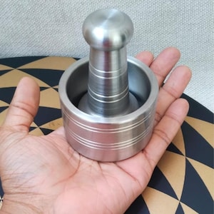 Puede incluir: Un juego de mortero y maja de acero inoxidable. El mortero es cilíndrico con líneas grabadas horizontales, y la maja tiene una parte superior redondeada. El juego está diseñado para moler hierbas y especias.