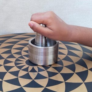 May include: A stainless steel mortar and pestle set. The mortar is cylindrical with horizontal grooves, and the pestle is held in place. The set is on a patterned surface.