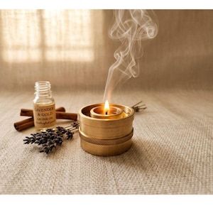 May include: A burning candle in a gold-coloured holder, with smoke rising. Next to it are a small bottle labelled "Lavender & Gold Aroma Oil", cinnamon sticks, and lavender sprigs. The items are on a textured, neutral-toned surface.