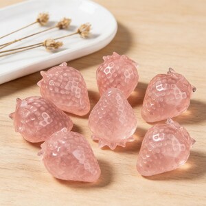 May include: A collection of pink strawberry-shaped crystals. The crystals are arranged on a wooden surface, with a white ceramic dish and dried flowers in the background. The strawberries have a textured surface.