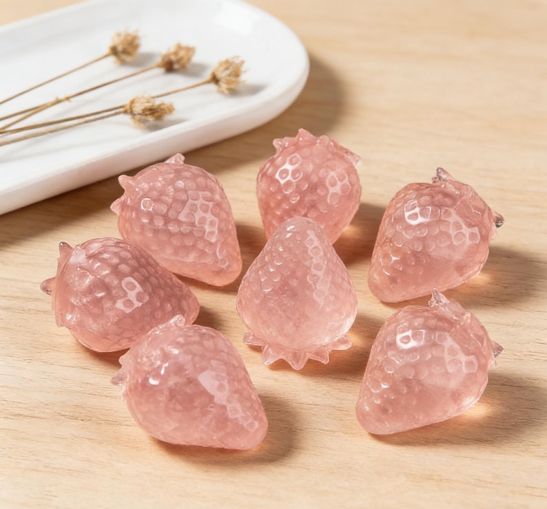 May include: A collection of pink strawberry-shaped crystals. The crystals are arranged on a wooden surface, with a white ceramic dish and dried flowers in the background. The strawberries have a textured surface.