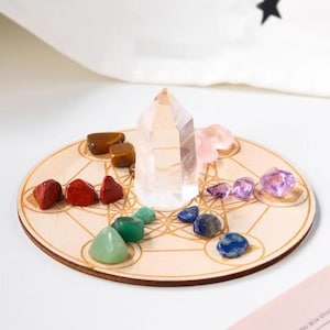 May include: A wooden crystal grid with a clear quartz point and an assortment of colorful gemstones. The gemstones include red, green, blue, purple, and brown stones. The grid has a geometric design.