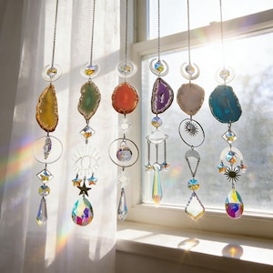 May include: A collection of hanging suncatchers with colorful agate slices and crystal accents. Each suncatcher features a unique design with metal charms, including stars, moons, and geometric shapes. The agate slices are in shades of yellow, green, orange, purple, and blue.