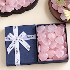 May include: A navy blue gift box with a white ribbon that reads "Crystal Jewelry" is open, revealing a collection of pink heart-shaped stones. Additional heart-shaped stones are displayed in wooden trays.