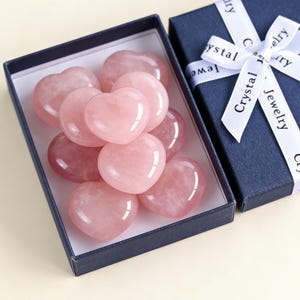 May include: A collection of polished rose quartz heart-shaped stones in a navy blue gift box. The box is partially open, revealing the pink hearts. A matching navy blue box with a white ribbon that reads "Crystal Jewelry" is next to it.