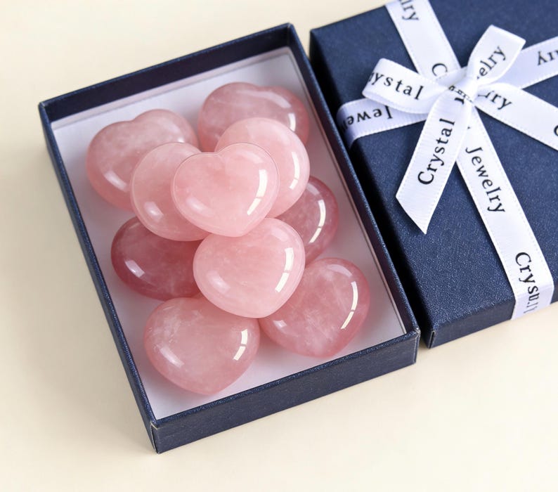 May include: A collection of polished rose quartz heart-shaped stones in a navy blue gift box. The box is partially open, revealing the pink hearts. A matching navy blue box with a white ribbon that reads "Crystal Jewelry" is next to it.