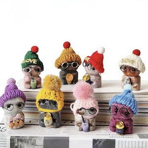 May include: A collection of whimsical figurines, each adorned with a knitted hat and round glasses. The figurines hold beverages and are painted in various colors, including green, yellow, pink, and blue. The hats have pom-poms.