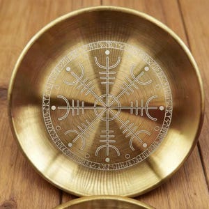 K&ouml;nnte beinhalten: Eine runde, messingfarbene Schale mit detailliertem, ge&auml;tztem Design. Das zentrale Motiv ist ein symmetrischer, stilisierter Kompass oder ein Rad, umgeben von einem Ring aus Runen. Die Schale hat eine leicht konkave Form und eine polierte Oberfl&auml;che.