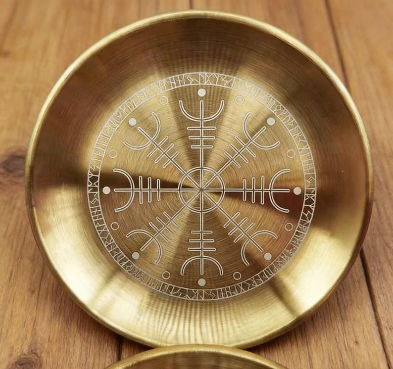 K&ouml;nnte beinhalten: Eine runde, messingfarbene Schale mit detailliertem, ge&auml;tztem Design. Das zentrale Motiv ist ein symmetrischer, stilisierter Kompass oder ein Rad, umgeben von einem Ring aus Runen. Die Schale hat eine leicht konkave Form und eine polierte Oberfl&auml;che.
