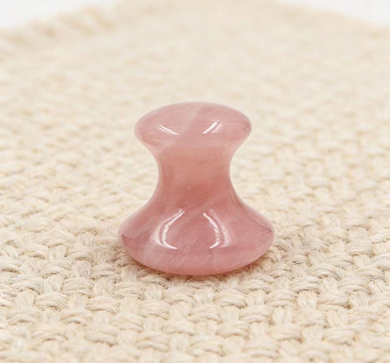 May include: A pink rose quartz facial massage tool. The tool has a rounded top, a narrow neck, and a wider base. The polished stone has a translucent appearance and is set against a neutral background.