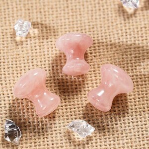 May include: Three polished rose quartz mushroom-shaped massage tools. The pink stones are smooth and glossy, with a rounded top and a flared base. Clear crystal fragments are scattered around the stones on a textured, tan surface.