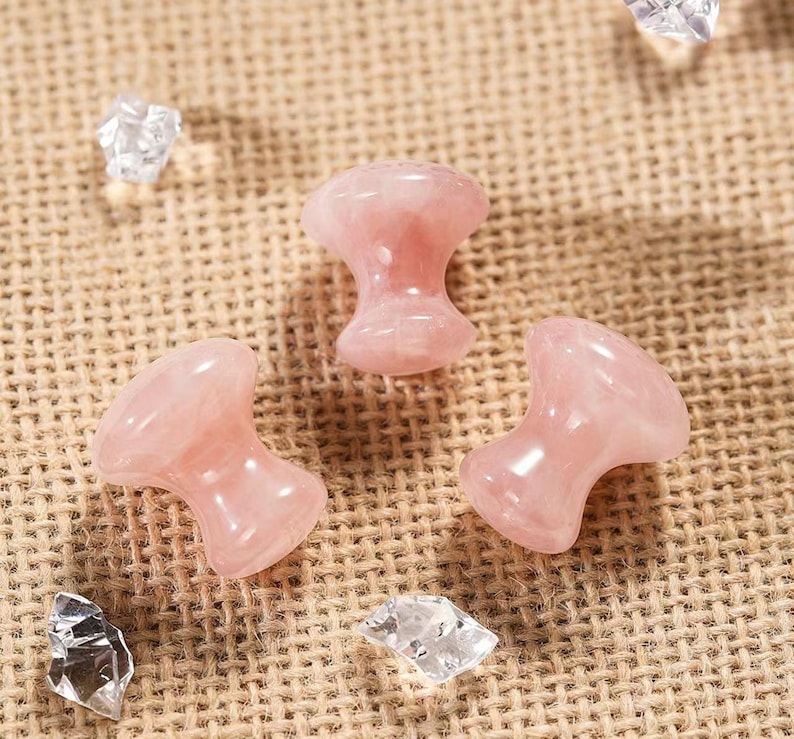 May include: Three polished rose quartz mushroom-shaped massage tools. The pink stones are smooth and glossy, with a rounded top and a flared base. Clear crystal fragments are scattered around the stones on a textured, tan surface.