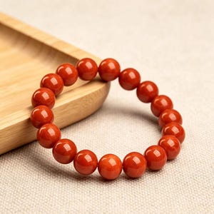 Könnte beinhalten: Ein elastisches Armband bestehend aus zahlreichen glatten, kugelförmigen Perlen in einer leuchtenden, einfarbigen roten Farbe. Die Perlen sind in Größe und Form einheitlich und kreisförmig angeordnet, um eine geschlossene Schlaufe zu bilden.