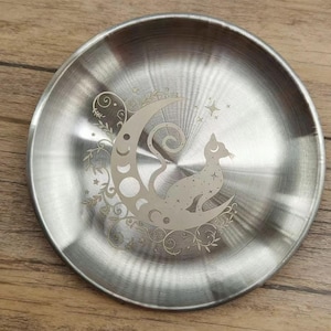 K&ouml;nnte beinhalten: Eine runde, silberfarbene Metallplatte mit einem ge&auml;tzten Design. Das Design zeigt eine Katze, die auf einem Mond sitzt, umgeben von Sternen und floralen Akzenten. Die Platte hat eine geb&uuml;rstete Metalloberfl&auml;che.
