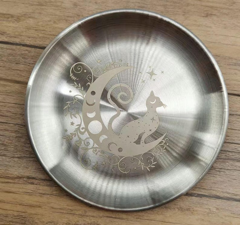 K&ouml;nnte beinhalten: Eine runde, silberfarbene Metallplatte mit einem ge&auml;tzten Design. Das Design zeigt eine Katze, die auf einem Mond sitzt, umgeben von Sternen und floralen Akzenten. Die Platte hat eine geb&uuml;rstete Metalloberfl&auml;che.