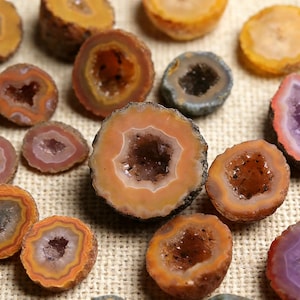 May include: A collection of sliced geodes in various colors, including orange, brown, purple, and gray. The geodes are cut to reveal the crystal formations inside, with a rough, textured exterior. The arrangement is on a neutral-toned, textured surface.