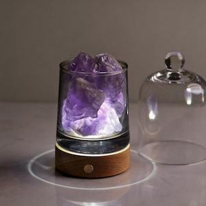 May include: A glass tumbler filled with purple amethyst crystals, illuminated from below by a soft white light. The tumbler sits on a wooden base with a single button. A clear glass cloche is next to the tumbler.