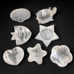 Peut inclure: Collection de sept plats décoratifs blancs translucides en forme de coquillage, tortue, poisson, étoile, cœur, étoile de mer et fleur. Les plats sont disposés sur une surface noire.