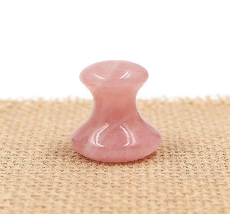 May include: A pink rose quartz facial massager with a rounded top and base, connected by a narrower neck. The smooth, polished stone is displayed on a textured, tan surface, against a white background. This beauty tool is designed for skincare.