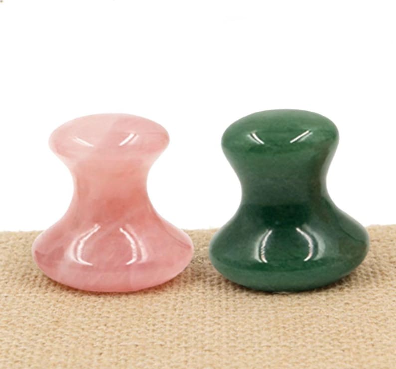 May include: Two gemstone facial massage tools. One is a light pink rose quartz, and the other is a deep green jade. Both have a rounded top and a flared base. They are displayed on a tan surface.