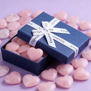 May include: A navy blue gift box tied with a white ribbon printed with "Crystal Jewelry" contains several pink heart-shaped stones. More heart-shaped stones are scattered around the box on a purple surface. The stones are a light pink color.