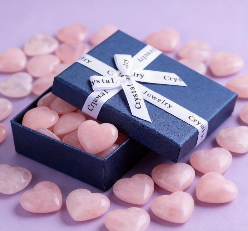 May include: A navy blue gift box tied with a white ribbon printed with "Crystal Jewelry" contains several pink heart-shaped stones. More heart-shaped stones are scattered around the box on a purple surface. The stones are a light pink color.