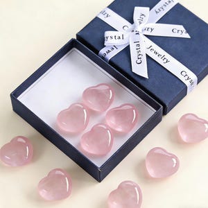 May include: A navy blue gift box with a white ribbon that reads "Crystal Jewelry" is open to reveal several pink heart-shaped crystals. Some hearts are inside the box, and others are scattered around it.