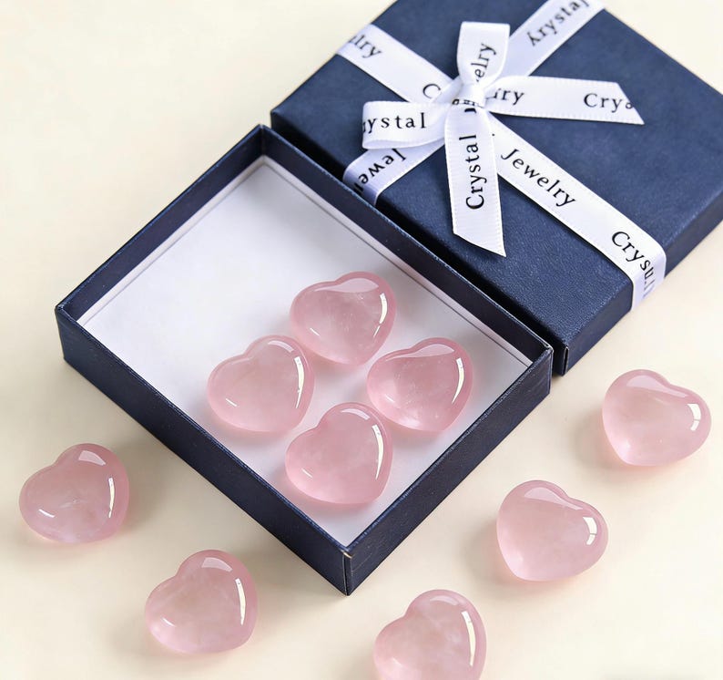 May include: A navy blue gift box with a white ribbon that reads "Crystal Jewelry" is open to reveal several pink heart-shaped crystals. Some hearts are inside the box, and others are scattered around it.