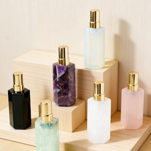 Può includere: Un assortimento di flaconi di profumo rettangolari con vaporizzatori color oro. I flaconi sono in vari colori, tra cui nero, viola, bianco, verde e rosa. Sono disposti su un espositore in legno, mostrando i loro design e colori unici.