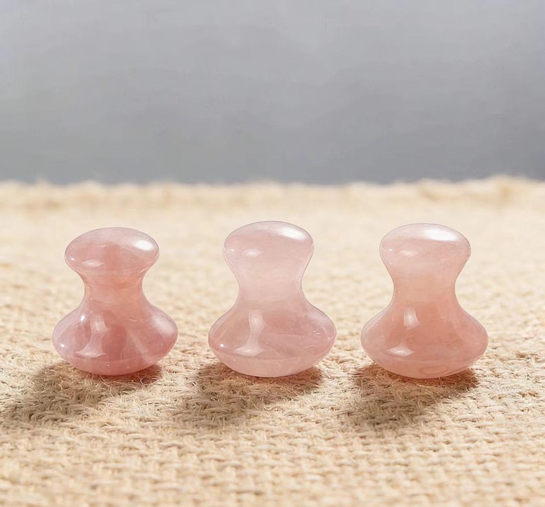 May include: Three rose quartz facial massage tools. Each tool has a rounded top and a flared base. The tools are a soft pink color and sit on a tan, textured surface. The background is a neutral gray.