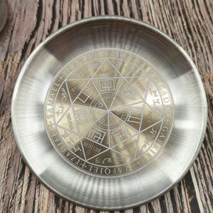 Könnte beinhalten: Ein runder, silberfarbener Metallteller mit einem geätzten goldenen Design in der Mitte. Das Design zeigt geometrische Formen und Text am Umfang. Der Teller liegt auf einer Holzoberfläche.