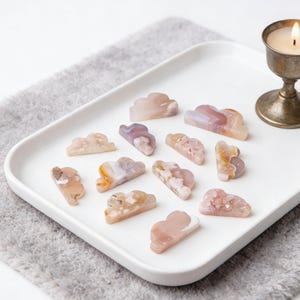 May include: A collection of cloud-shaped stones in shades of pink, purple, and cream, arranged on a white tray. A lit candle in a silver holder sits nearby. The stones have a polished, translucent appearance.