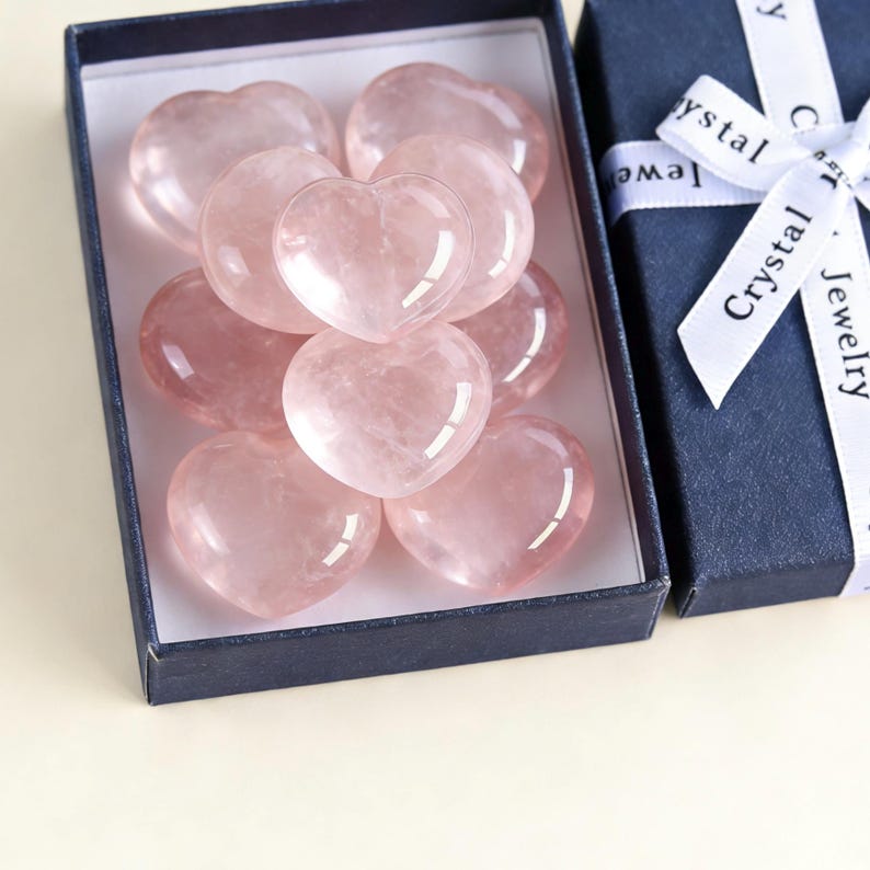 May include: A collection of pink rose quartz heart-shaped stones displayed in a navy blue gift box. The box is open, revealing the polished stones. A white ribbon with the words "Crystal Jewelry" is tied around the box.