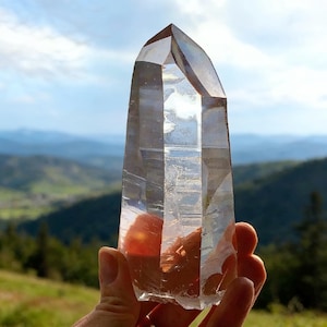 Puede incluir: Un cristal de cuarzo transparente, sostenido en una mano, con un fondo montañoso. El cristal es transparente, con estructuras internas y facetas visibles. El fondo muestra colinas verdes y un cielo azul.