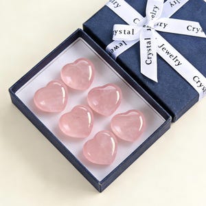 May include: A navy blue gift box with a white ribbon that reads "Crystal Jewelry" contains six polished rose quartz heart-shaped stones. The stones are a soft pink color and are arranged inside a white box.