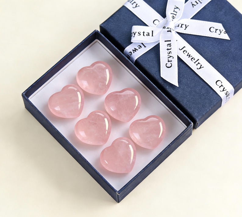 May include: A navy blue gift box with a white ribbon that reads "Crystal Jewelry" contains six polished rose quartz heart-shaped stones. The stones are a soft pink color and are arranged inside a white box.