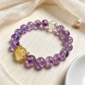 May include: A beaded bracelet featuring translucent purple amethyst spheres, one clear quartz bead, and one larger yellow citrine bead accented by gold-toned metal filigree caps. The beads are arranged in a circular pattern on a flexible cord.