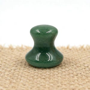 May include: A green jade massage tool with a rounded top and base, connected by a narrower midsection. The polished stone reflects light, and the tool sits on a textured, tan surface. The shape is ergonomic and designed for massage.